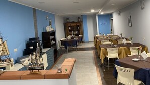 A dining area with tables set for a meal, a bookshelf, and a model ship on the left. - Kassiopea Aparthotel (Giardini Naxos)
