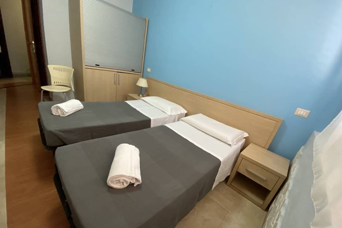 Een hotelkamer met twee eenpersoonsbedden, elk met een kussen en een opgerolde handdoek. Er is een houten hoofdbord, een nachtkastje met een lamp en een raam met jaloezieën.