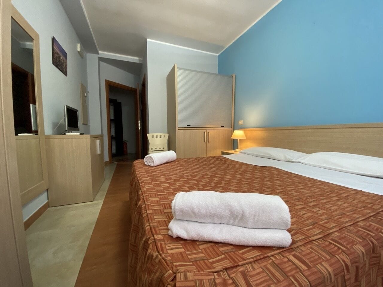 Een hotelkamer met een groot bed, een houten hoofdbord, een nachtkastje met een lamp en een bureau met een computer.