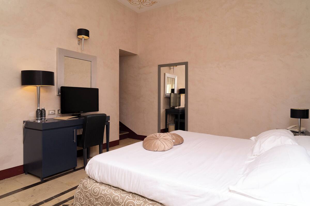 A hotel room with a bed, a desk with a computer, a chair, a desk lamp, a TV, and a mirror.