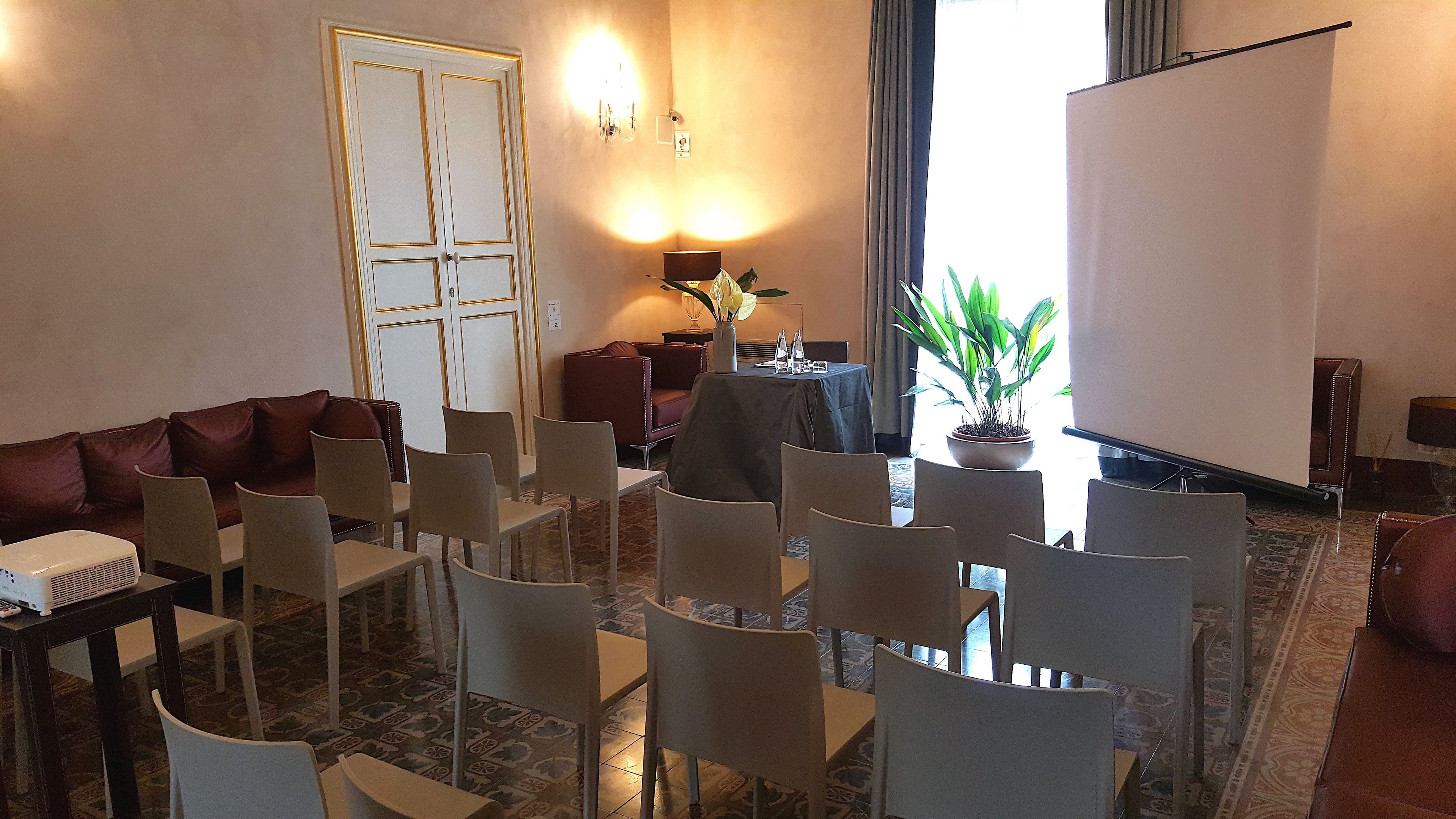 Ein Konferenzraum mit einer braunen Ledersitzgruppe, weißen Stühlen, einem Tisch mit Projektor, einer Topfpflanze und einer großen Whiteboard-Wand.