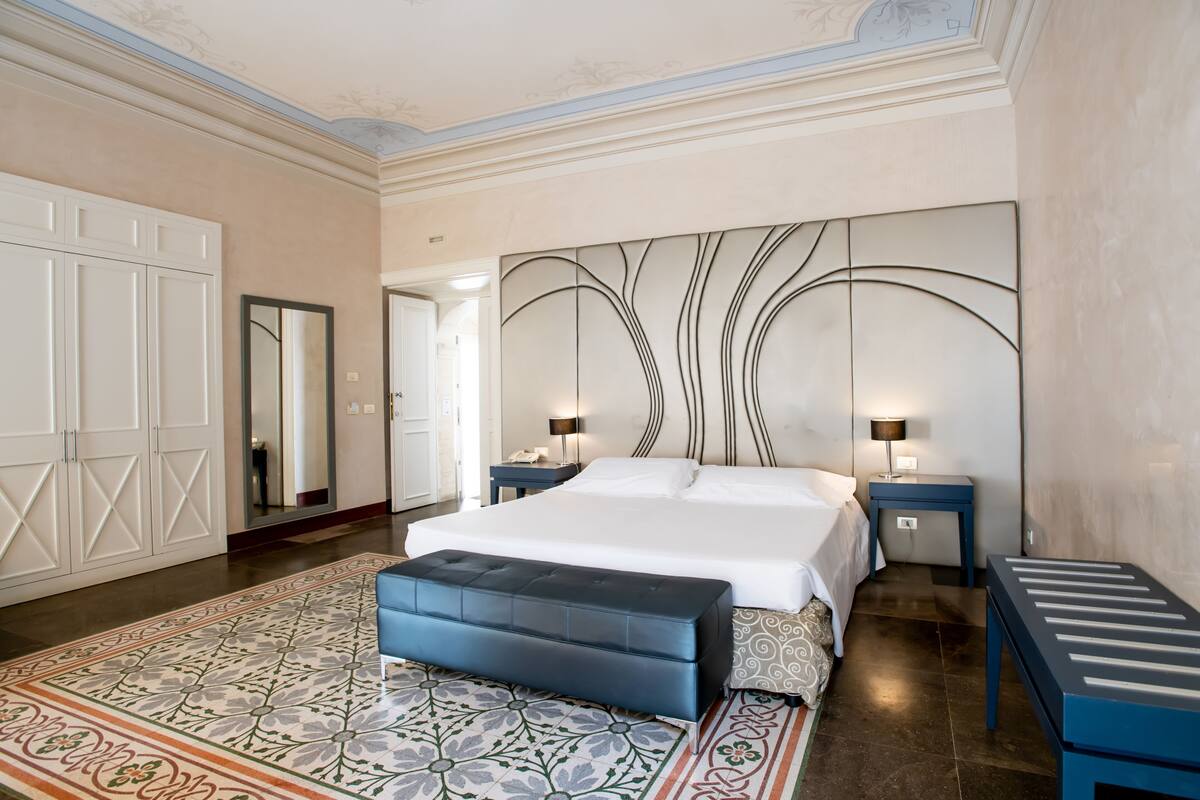 A bedroom with a large bed, a nightstand, a bench, and a decorative wall panel.