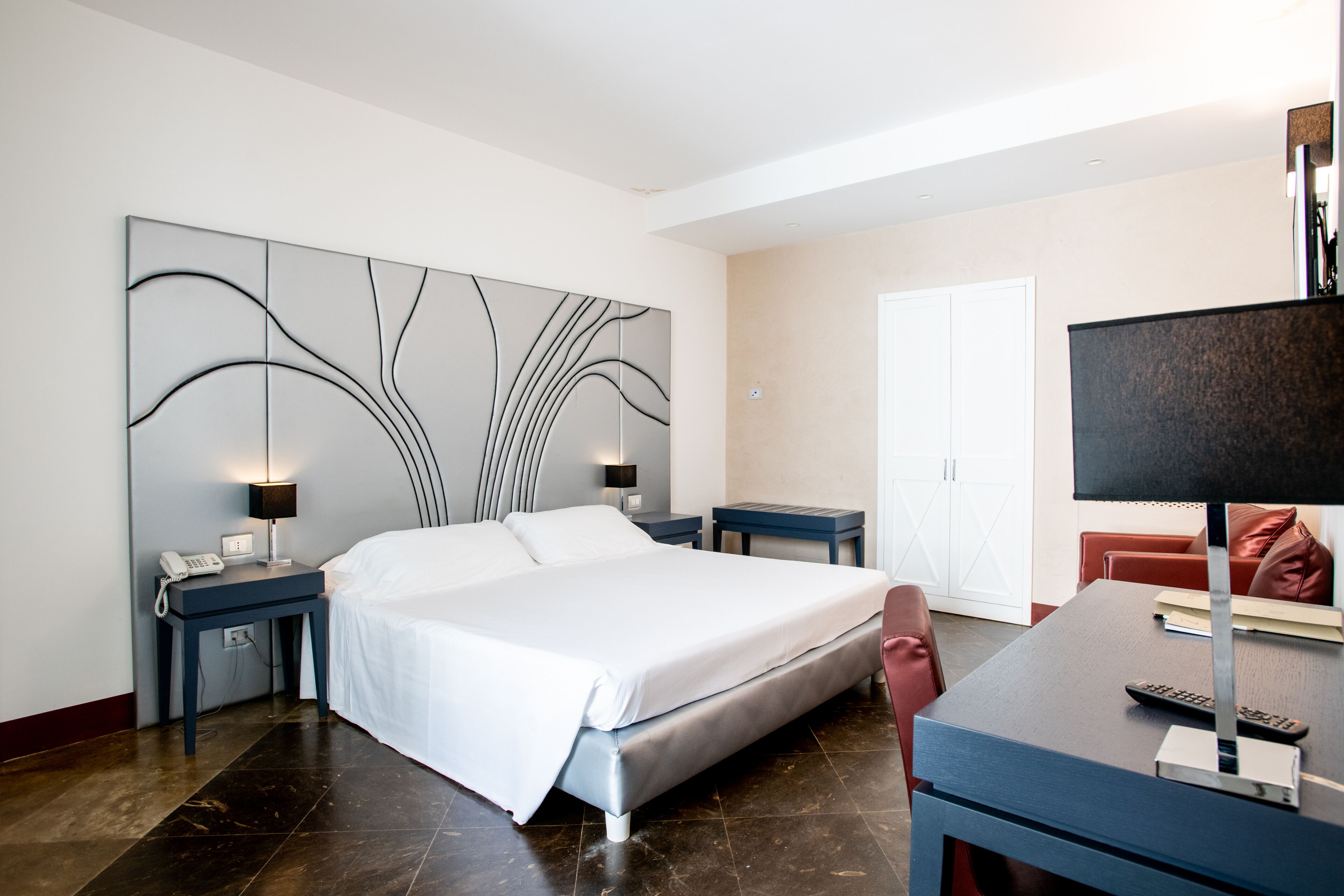 Una camera d'albergo moderna con un letto grande, una scrivania con un computer, una sedia, un comodino con una lampada e un'opera d'arte a parete.