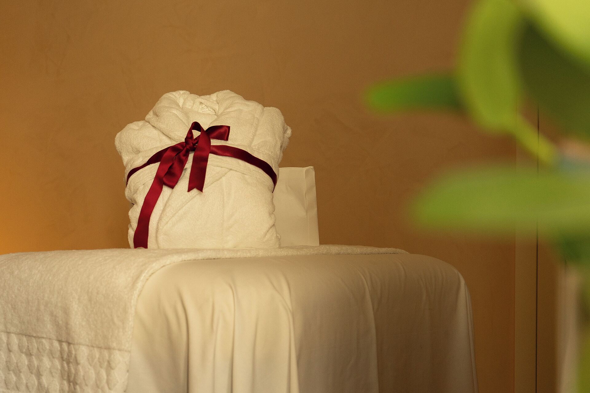 Une serviette blanche, soigneusement pliée et ornée d’un ruban rouge, est posée sur un lit recouvert d’une couverture blanche.
