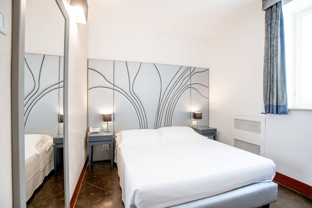 A modern hotel room with a large bed, bedside tables with lamps, a decorative wall panel, and a window with curtains.