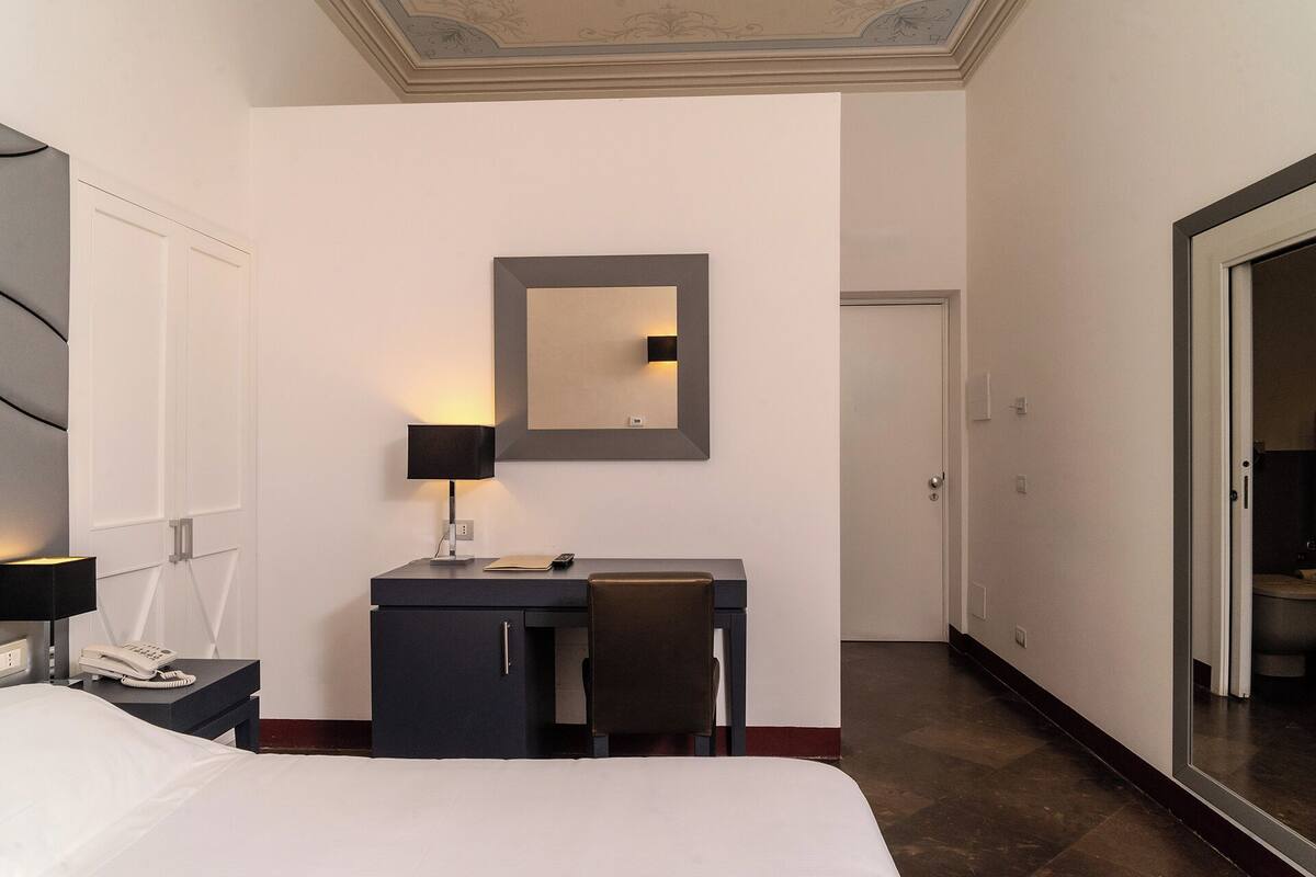 A hotel room with a bed, a desk with a chair, a lamp, a telephone, and a mirror.