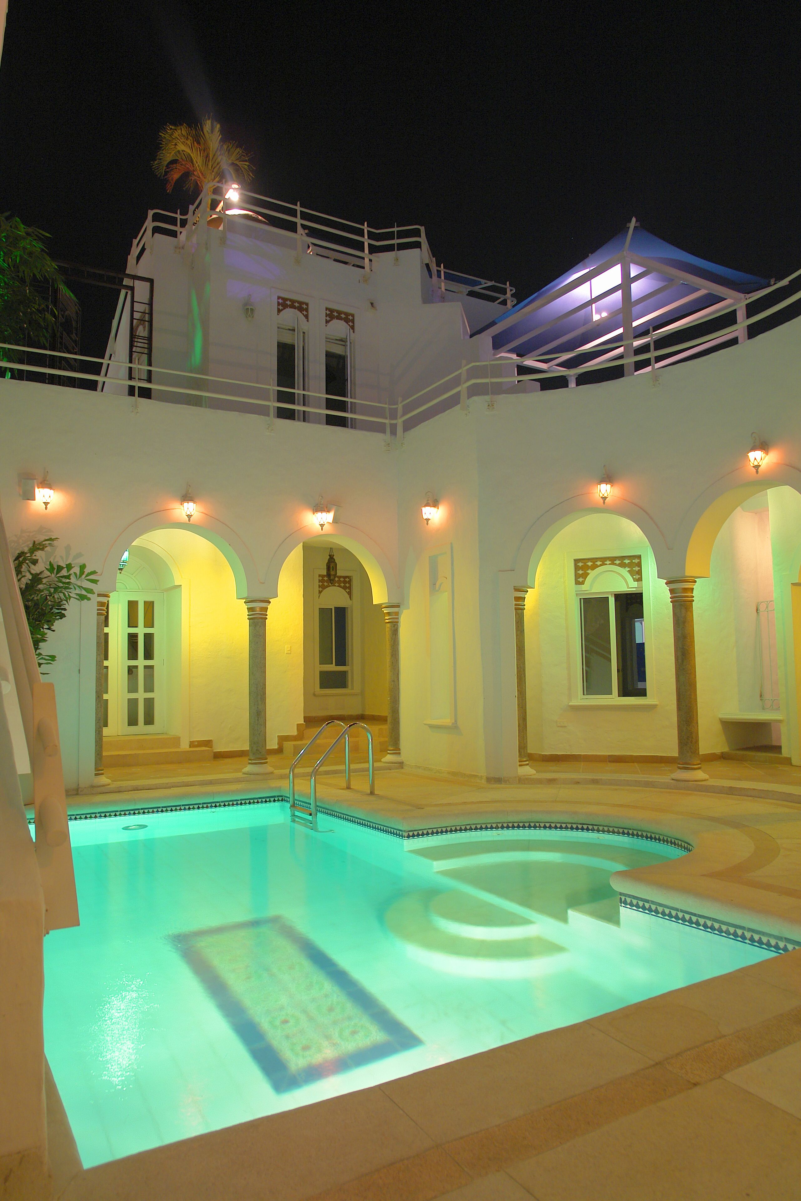 Ein Pool mit geometrischem Muster, umgeben von einem weißen Gebäude mit Rundbogenöffnungen und Säulen, sowie ein Balkon mit blauem Markisenvorzelt.