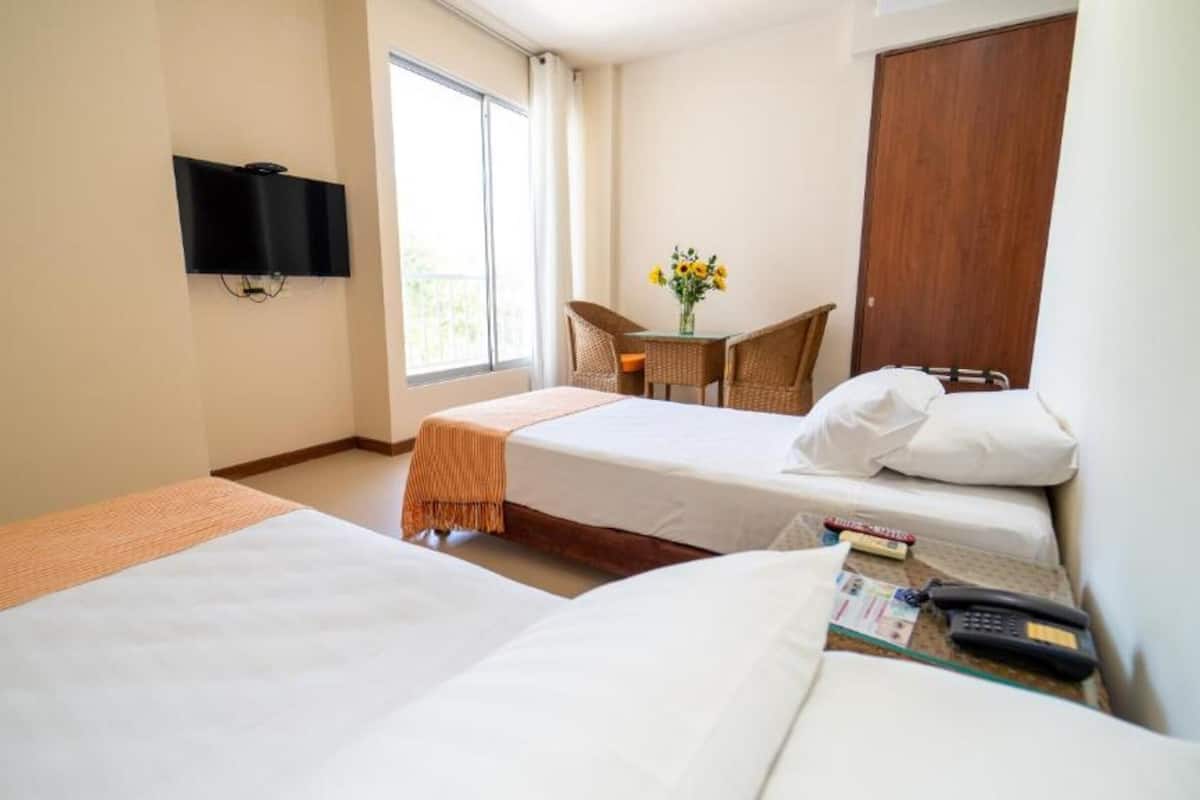 Quarto de hotel com duas camas, uma televisão presa na parede, uma mesinha com uma cadeira e uma janela com cortinas.
