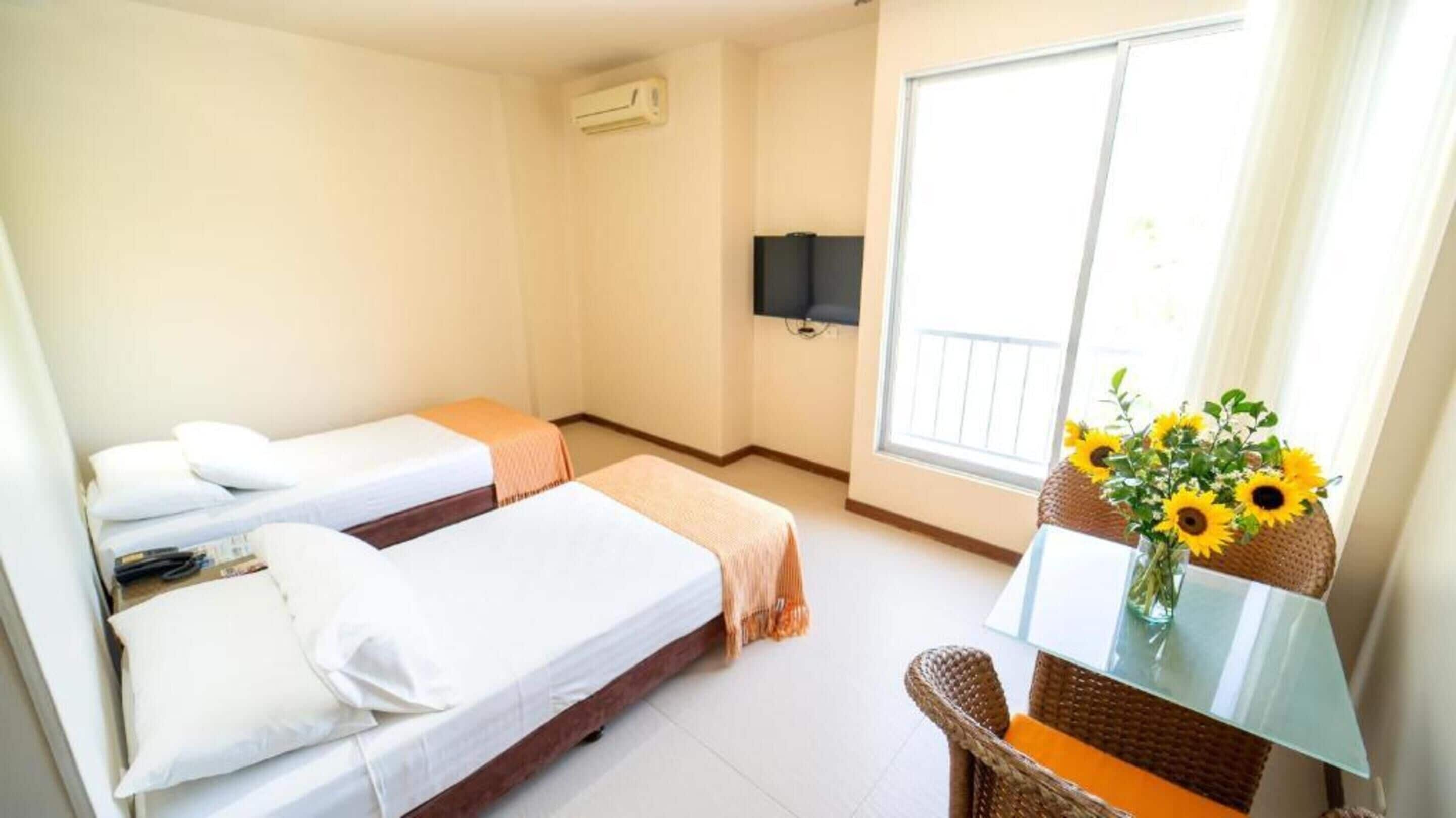 A hotel room with two single beds, a TV mounted on the wall, a window with a view, and a table with a vase of sunflowers.
