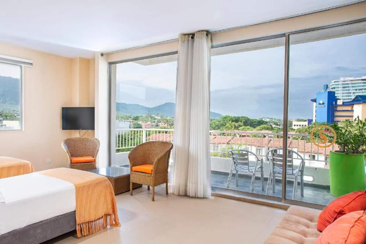Quarto de hotel com cama, poltrona, televisão e varanda com vista para a cidade e montanhas.