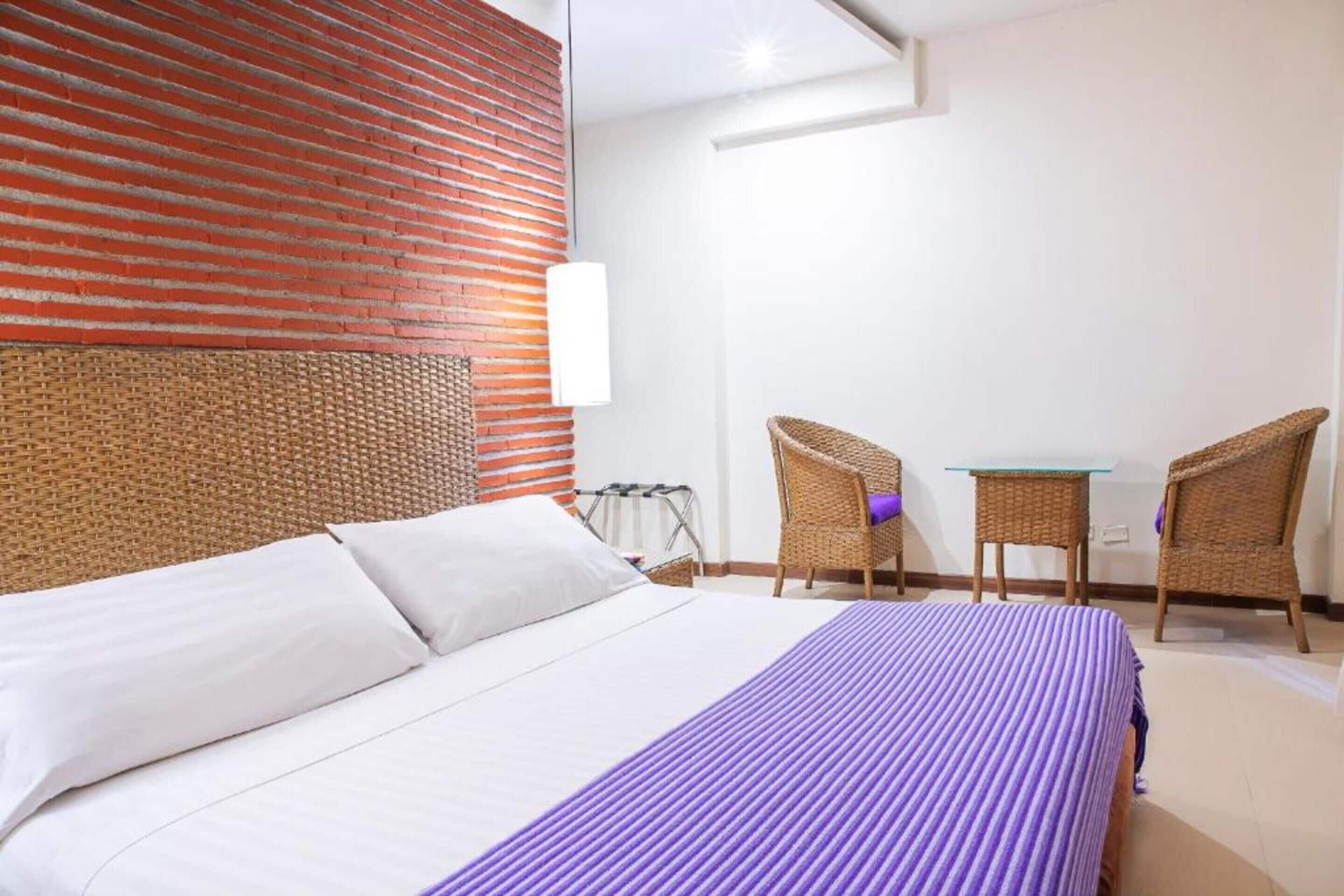 A hotel room with a bed, two wicker chairs, a small table, and a red brick wall.