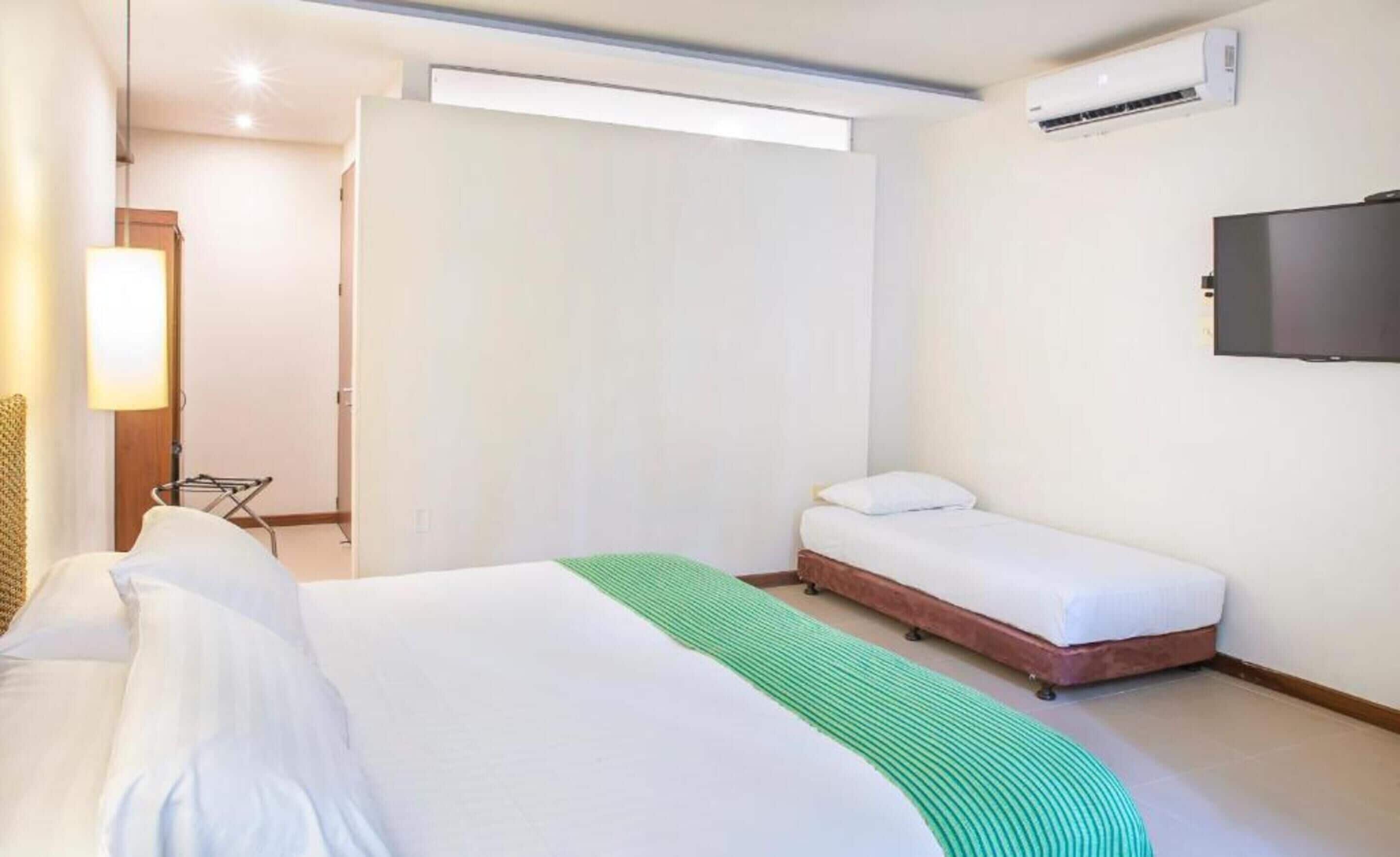 A hotel room with a bed, a green throw, a flat-screen TV, and an air conditioning unit.