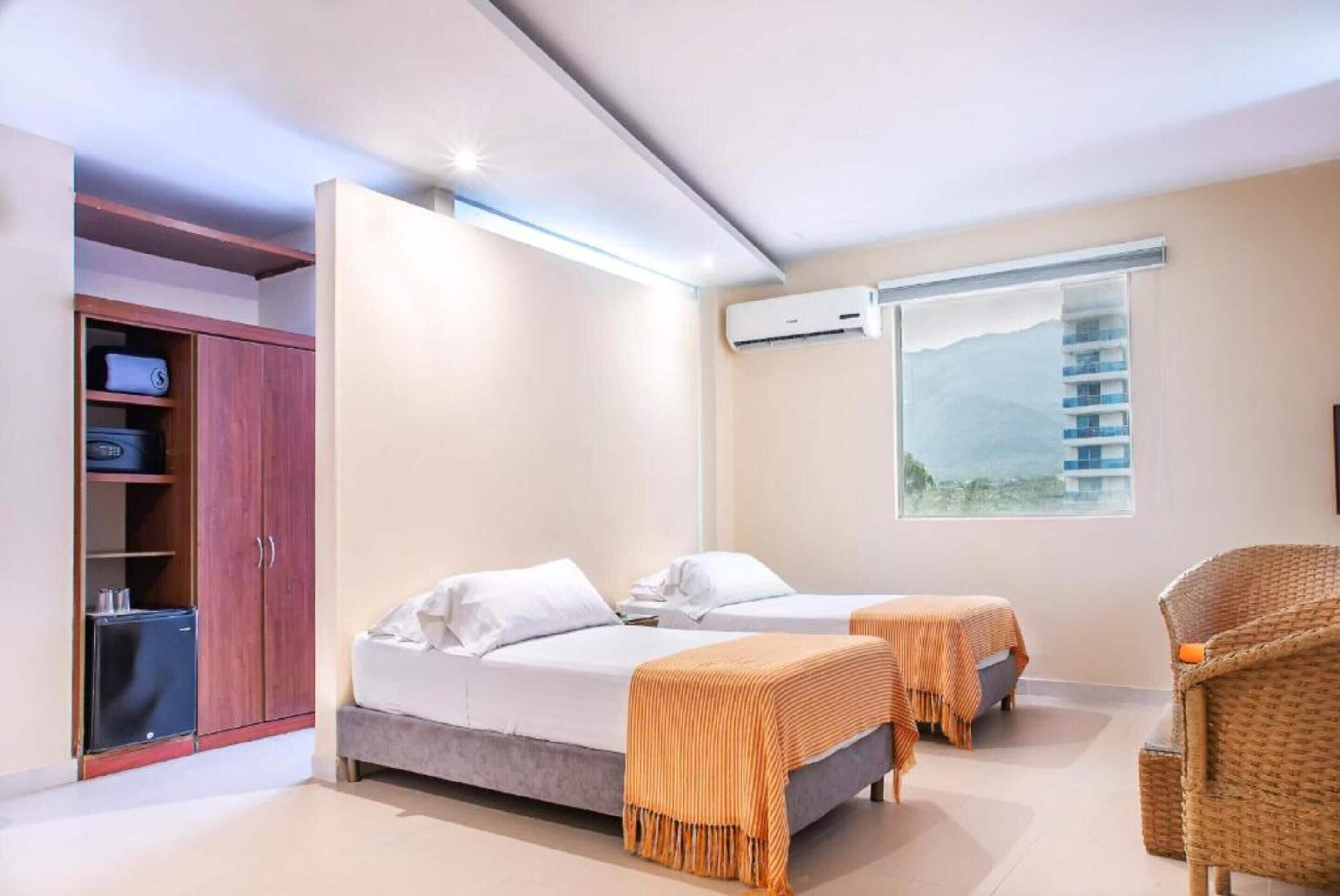 Ett modernt hotellrum med två sängar, ett litet bord, en flätad stol och en garderob.