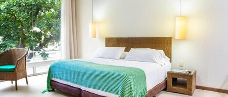 Habitación de hotel con una cama grande, una silla de mimbre, una mesita auxiliar y vistas a árboles por la ventana.