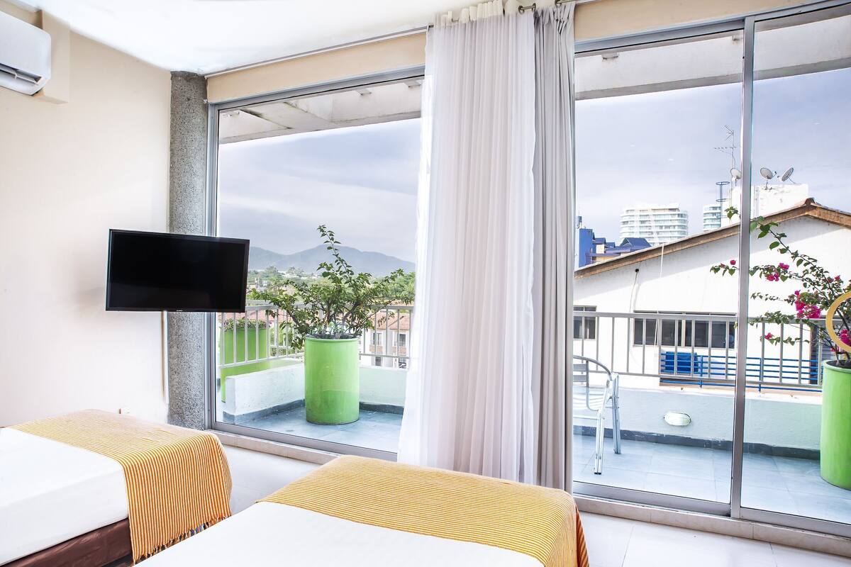 Quarto de hotel com duas camas, televisão, janelas amplas e varanda com plantas em vasos.