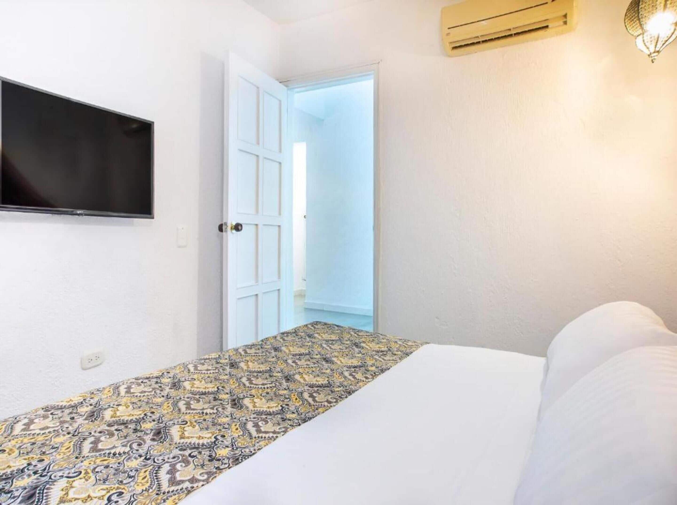 A bedroom with a bed, a TV mounted on the wall, a patterned bedspread, a door leading to a bright room, and an air conditioning unit.