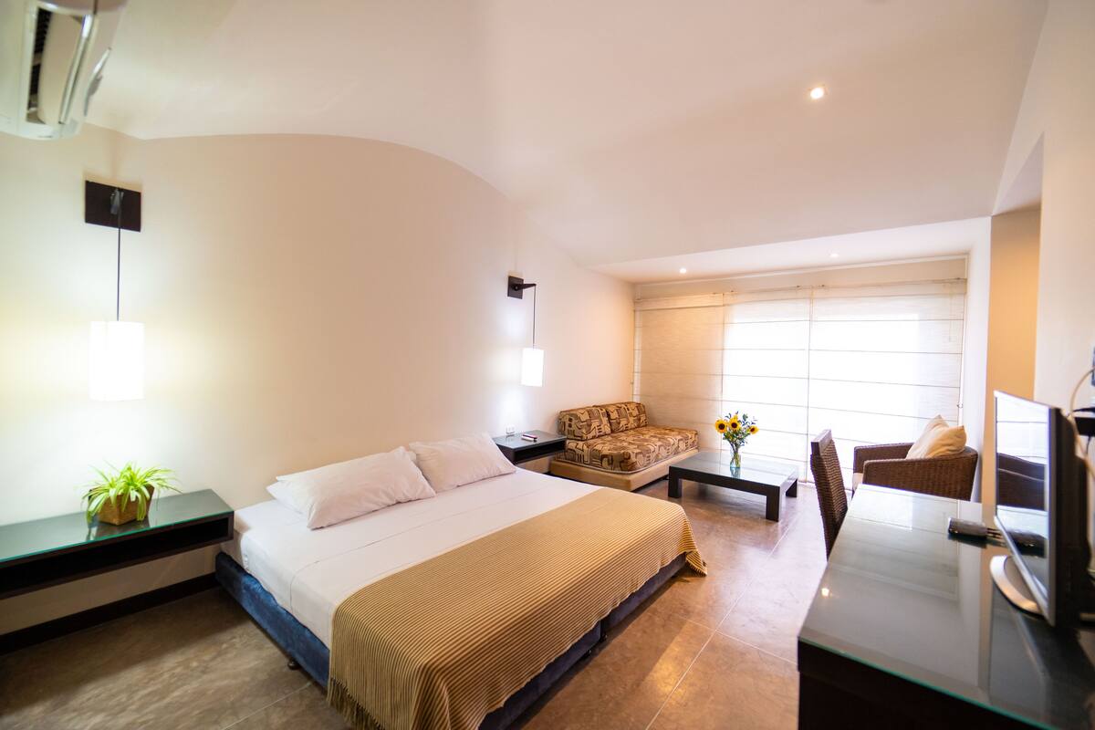 Une chambre d’hôtel moderne, dotée d’un grand lit, d’un canapé, d’un bureau avec un ordinateur et d’une fenêtre avec des stores.