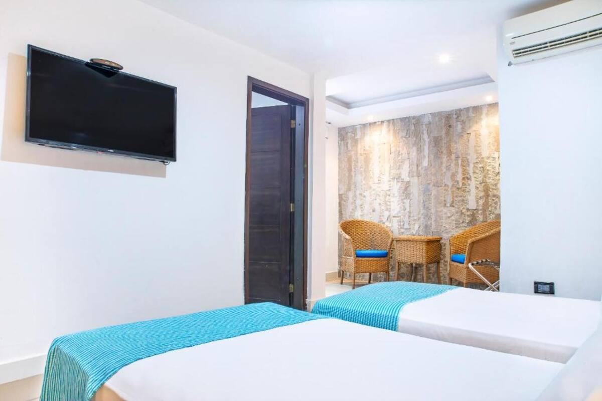 Une chambre d’hôtel avec deux lits, un téléviseur à écran plat, des fauteuils en osier et un mur à la texture de pierre.