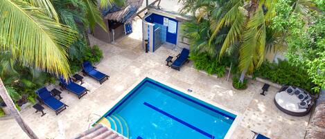 A swimming pool with lounge chairs and a hot tub, surrounded by palm trees and a thatched-roof structure.