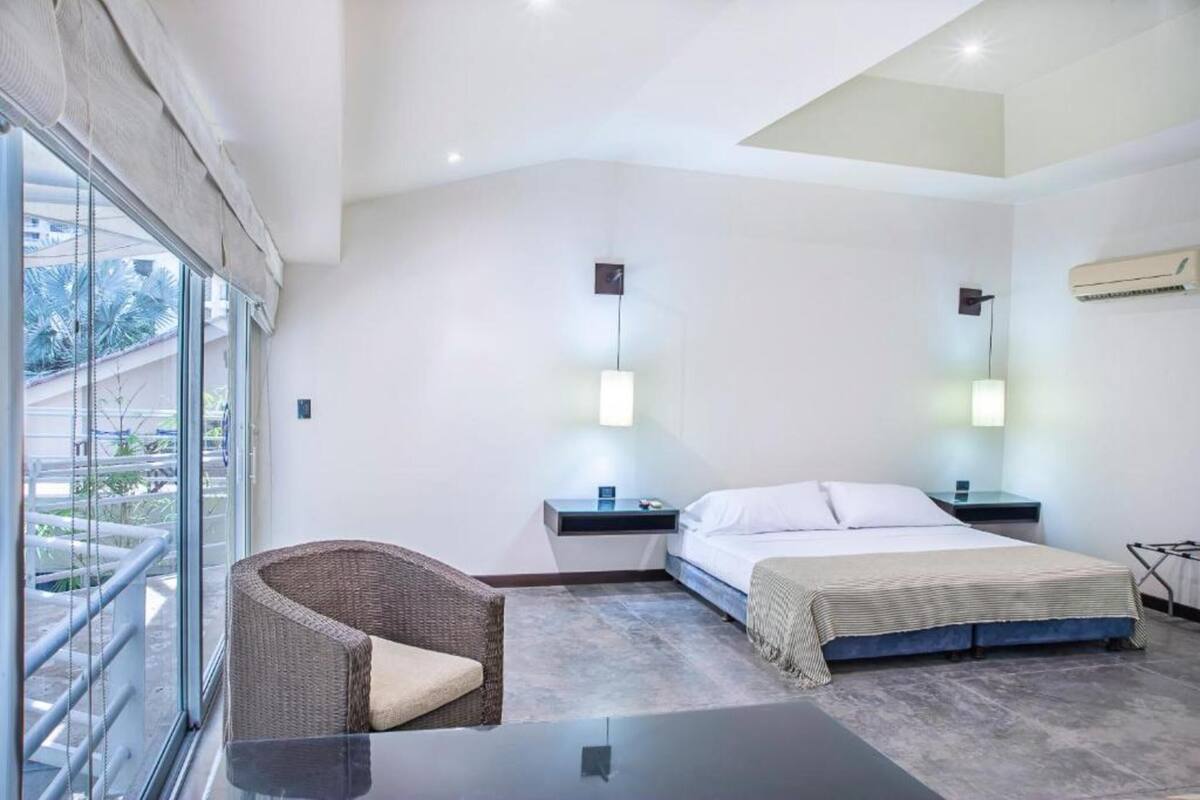 Une chambre d’hôtel moderne, équipée d’un lit, d’une chaise, d’une table de chevet et d’une grande fenêtre donnant sur l’extérieur.