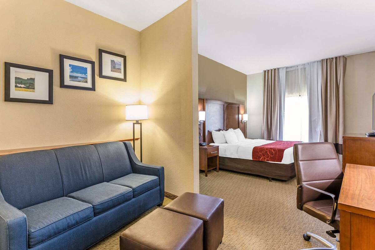 A hotel room with a blue sofa, a wooden desk, a bed with a red blanket, and framed pictures on the wall.