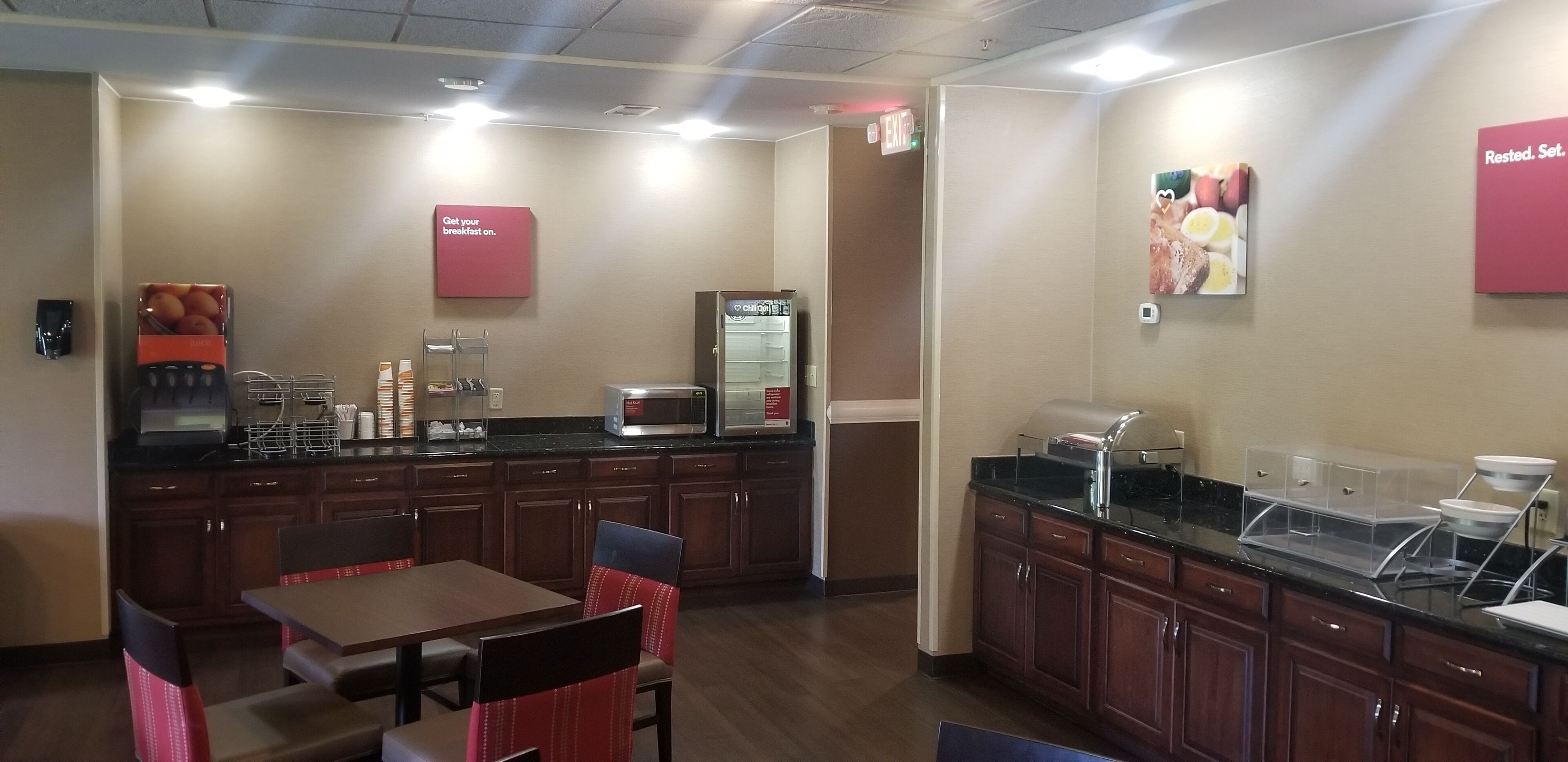 A hotel breakfast area with wooden cabinets, a microwave, a refrigerator, and a table with chairs.