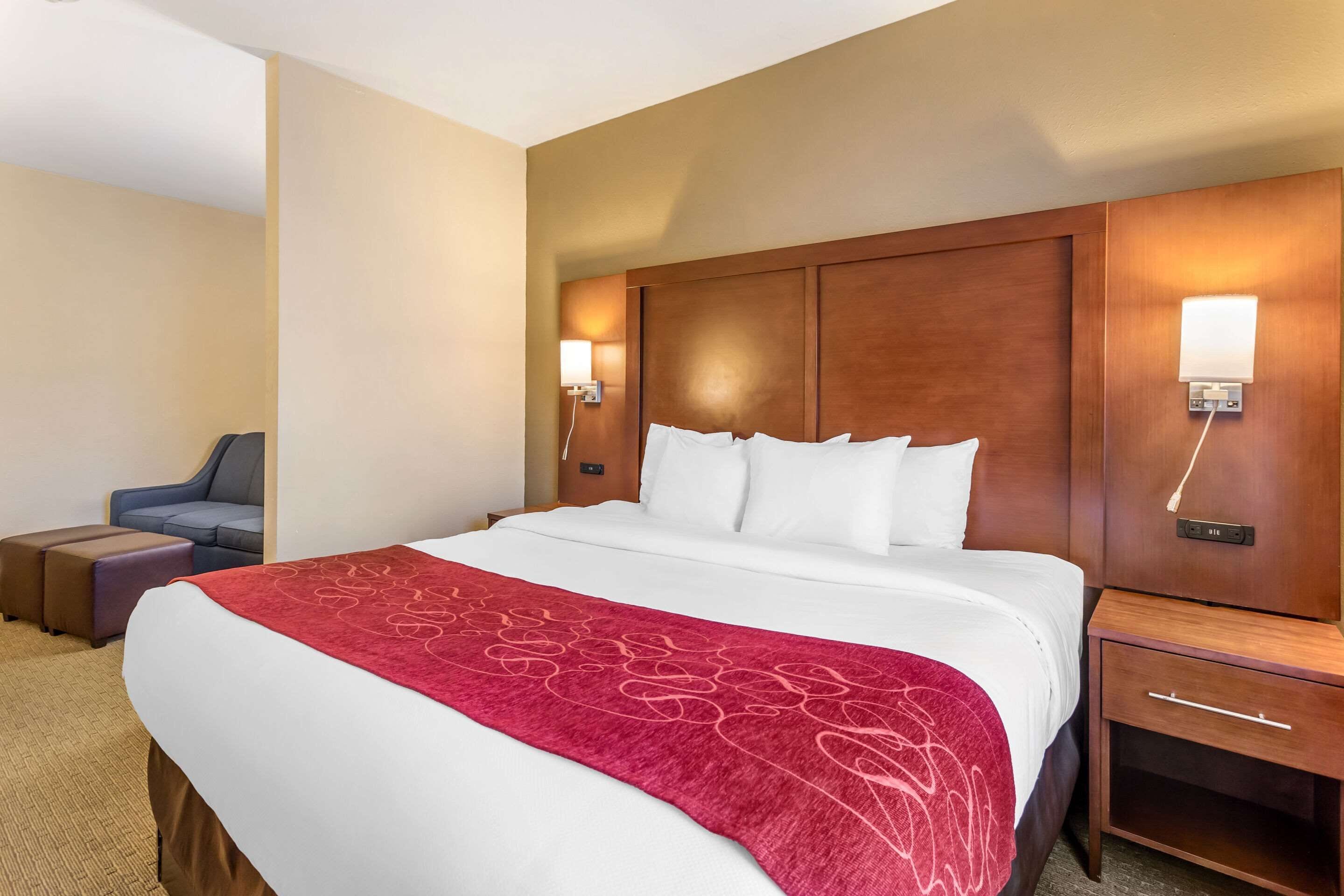 Hotelkamer met een groot bed, een houten hoofdeinde, twee nachtkastjes met lampen, een rode plaid en een grijze fauteuil.