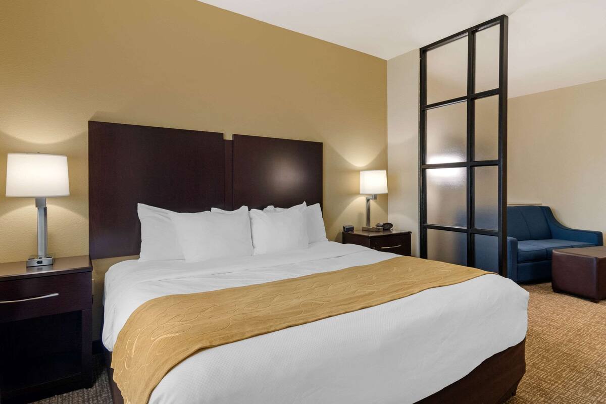 Une chambre d’hôtel avec un grand lit, deux tables de chevet avec des lampes, un fauteuil bleu et un grand miroir.