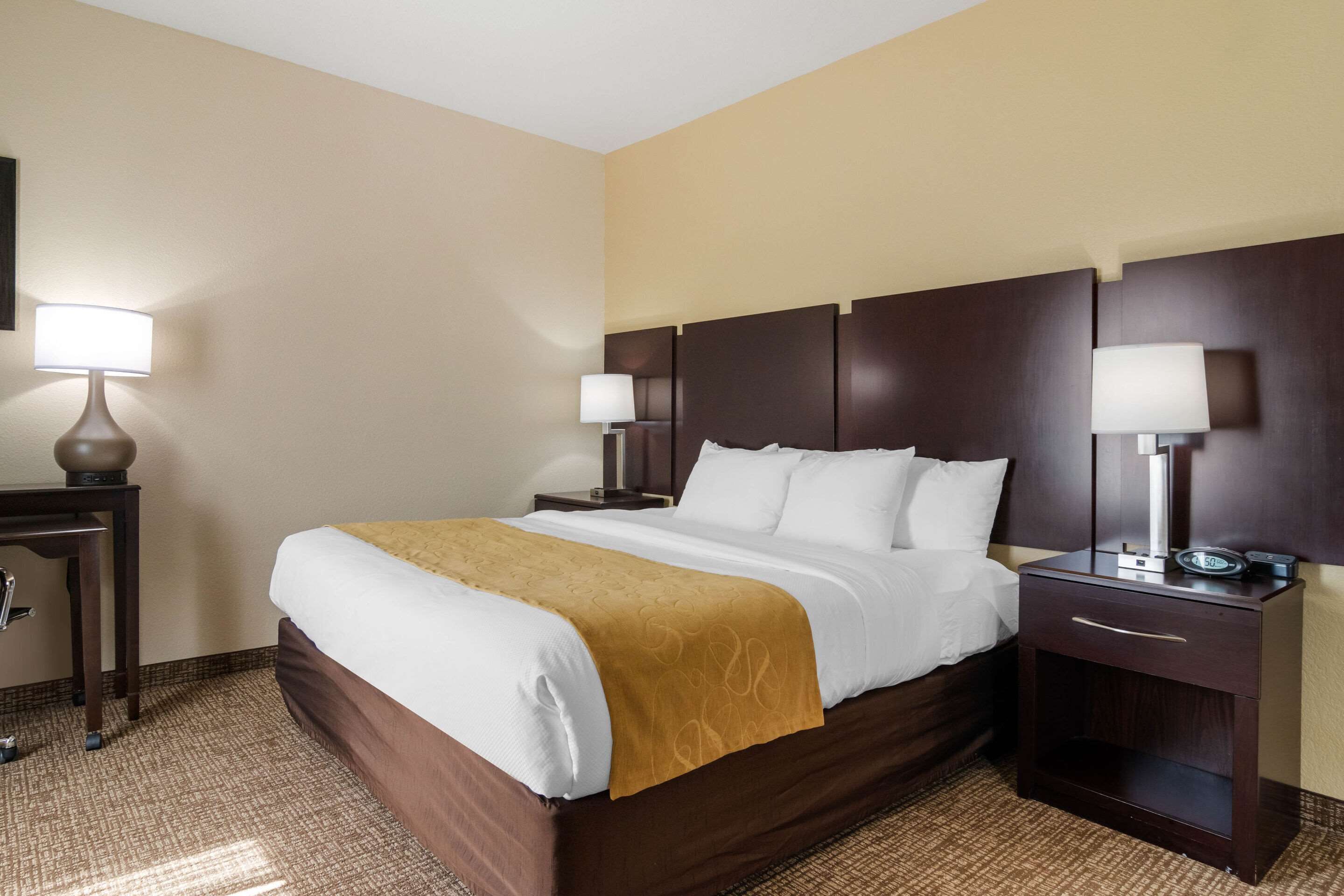 Quarto de hotel com uma cama grande, duas mesas de cabeceira com luminárias, uma cabeceira de madeira e piso coberto por carpete.