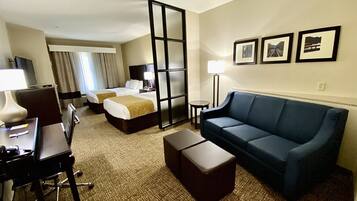 A hotel room with two beds, a sofa, a desk with a chair, a lamp, and framed pictures on the wall.