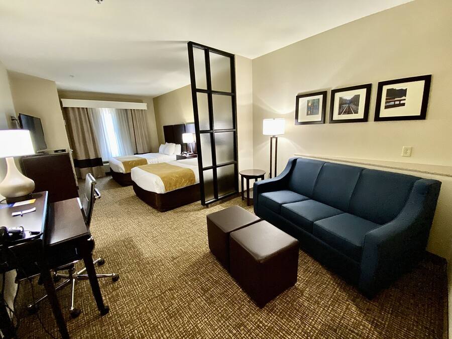 A hotel room with two beds, a sofa, a desk with a chair, a lamp, and framed pictures on the wall.