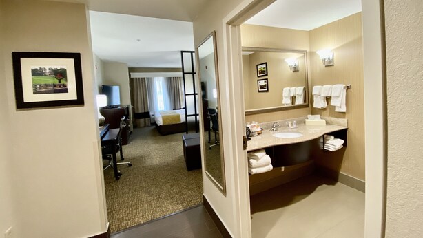 Habitación de hotel con cama, escritorio, silla, televisor y baño con lavamanos, espejo y toallas.