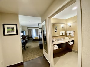 Habitación de hotel con cama, escritorio, silla, televisor y baño con lavamanos, espejo y toallas.