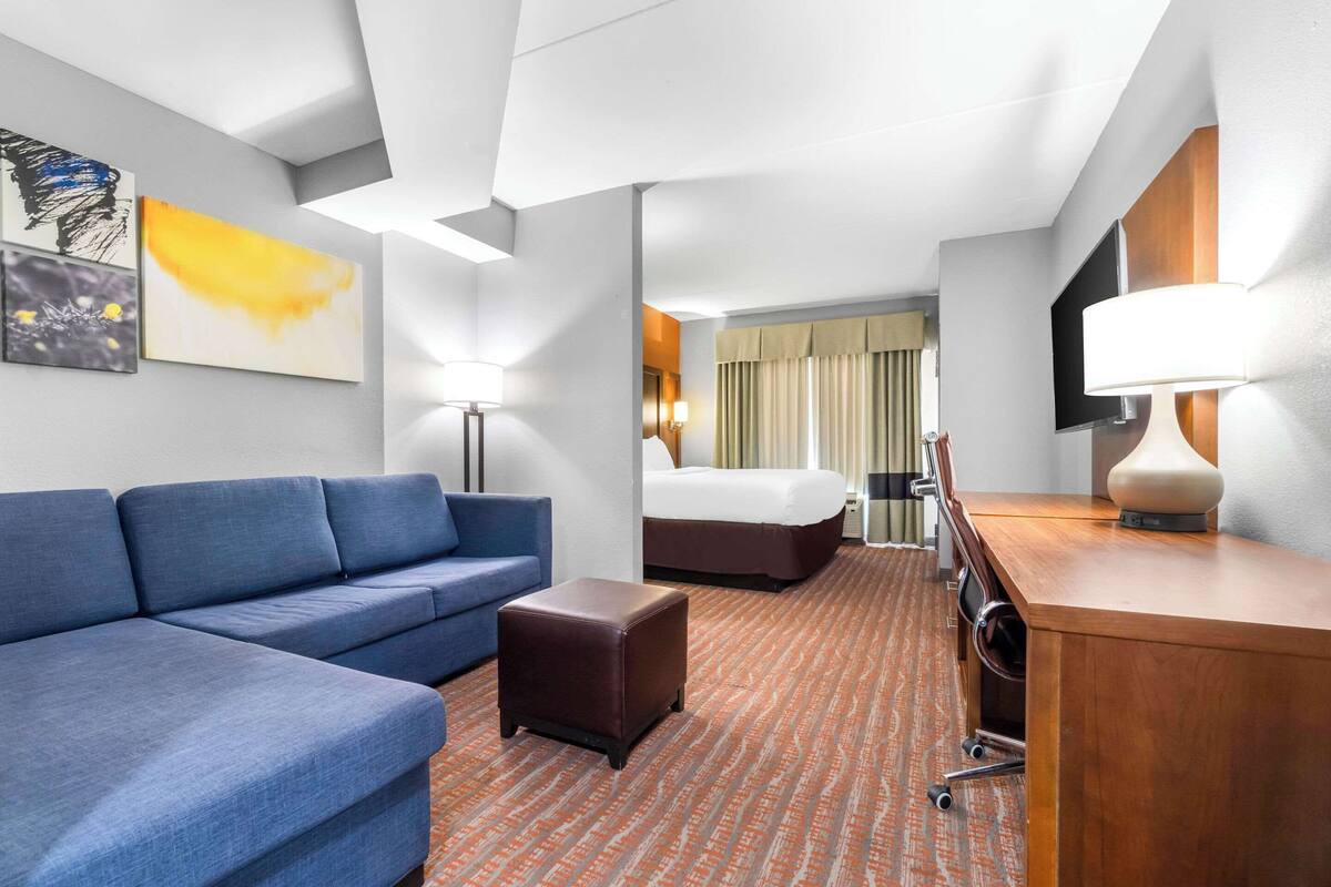 A hotel room with a blue sofa, a brown ottoman, a wooden desk, a bed with a beige headboard, and a flat-screen TV mounted on the wall.