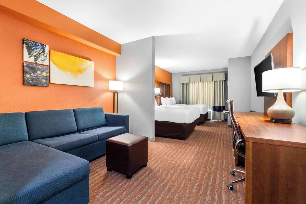 A hotel room with a sofa, a small table, a bed, a desk with a lamp, and artwork on the wall.