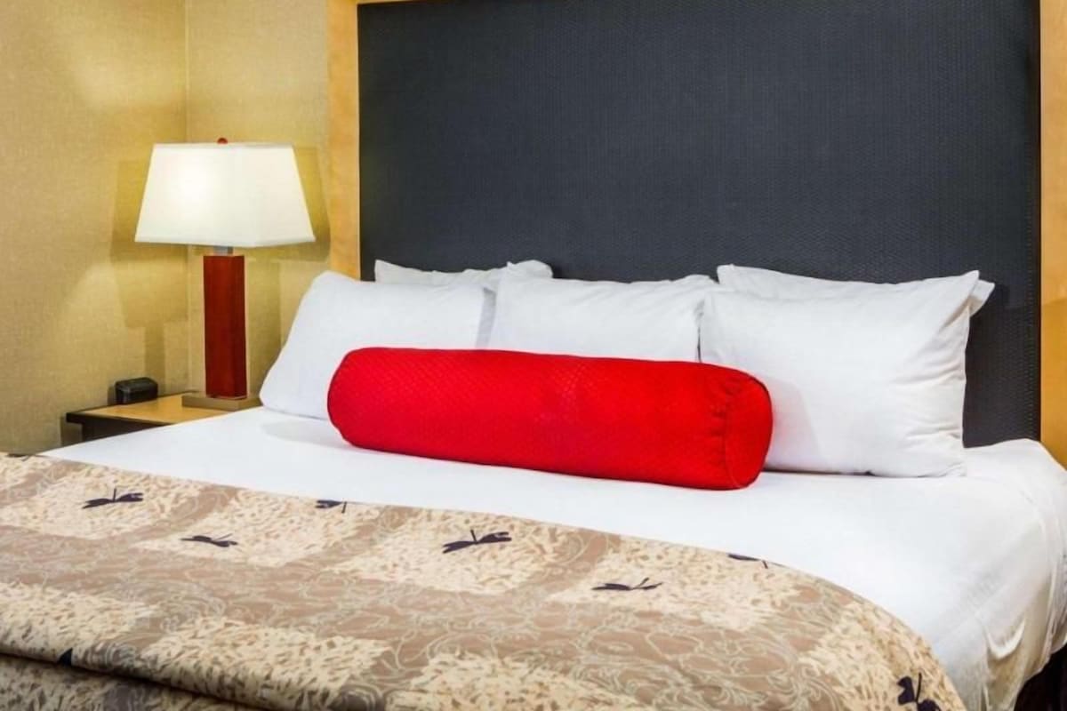 A hotel room with a bed, a nightstand, a lamp, and a red accent pillow.