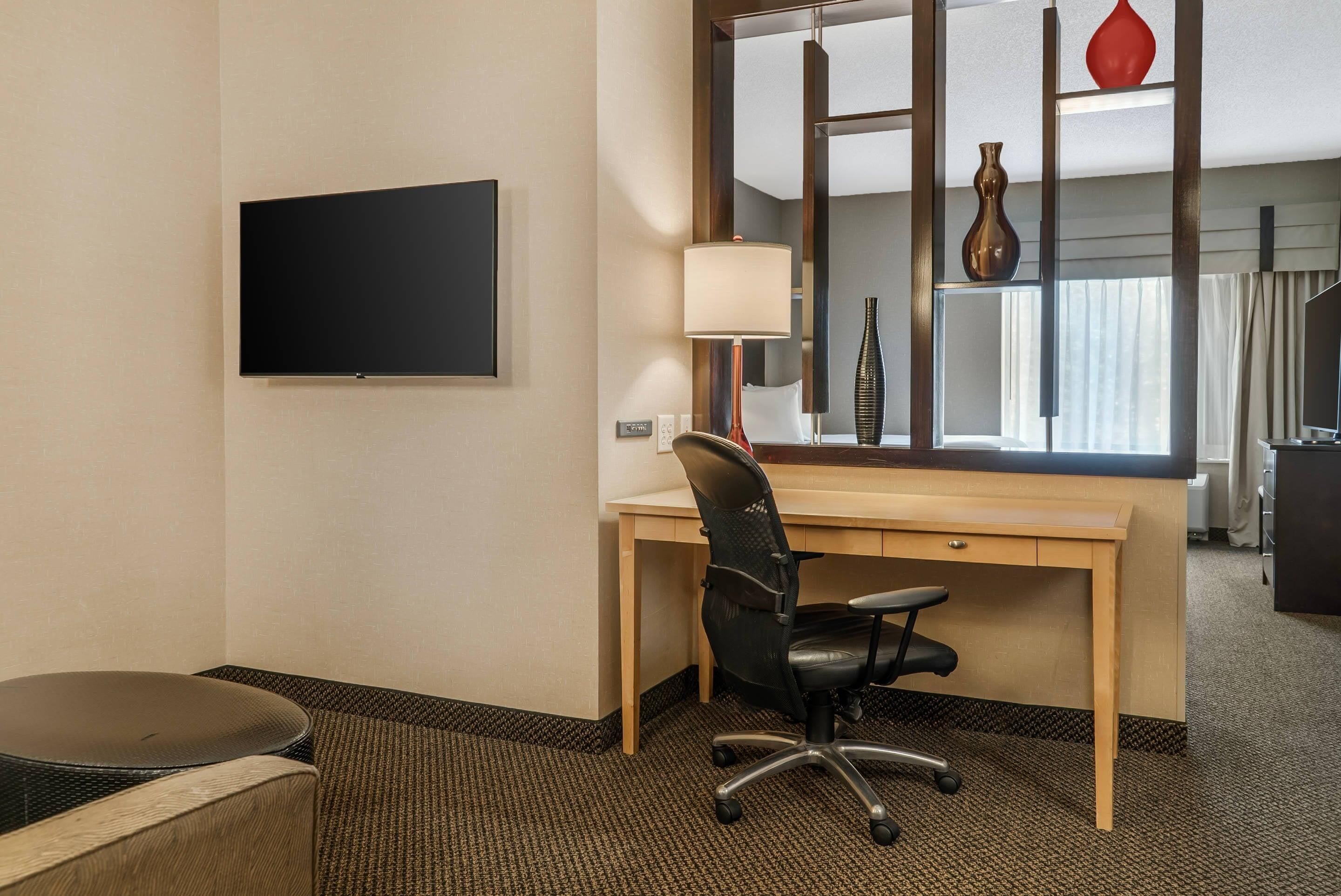Ett hotellrum med en platt-TV, ett skrivbord med en stol, en lampa och en stor spegel som reflekterar en vas och gardiner.