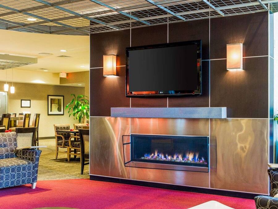 A modern lobby with a flat-screen TV mounted above a fireplace, comfortable seating, and a dining area in the background.
