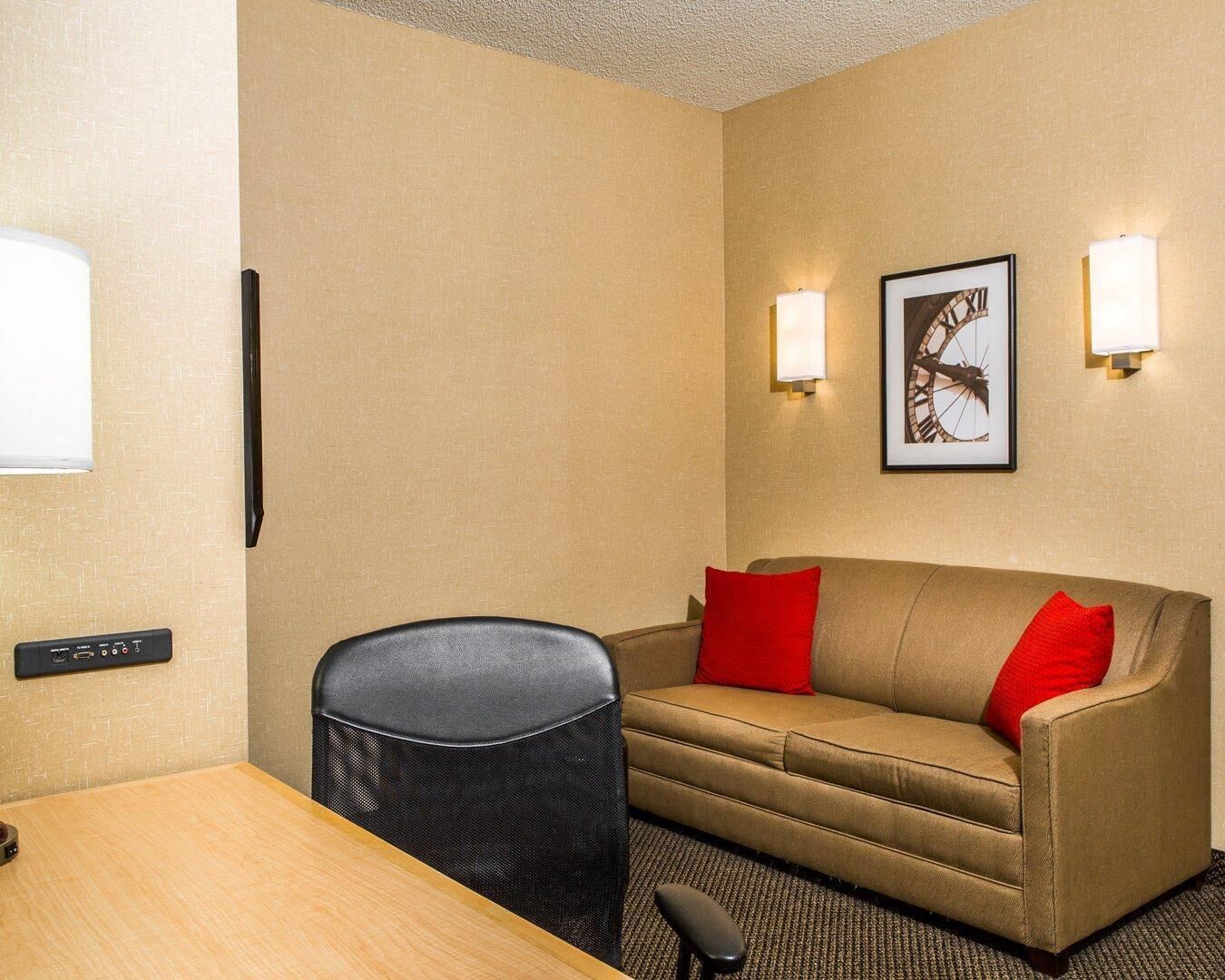 A hotel room with a beige sofa, two red cushions, a black chair, a wooden table, and a wall-mounted TV.