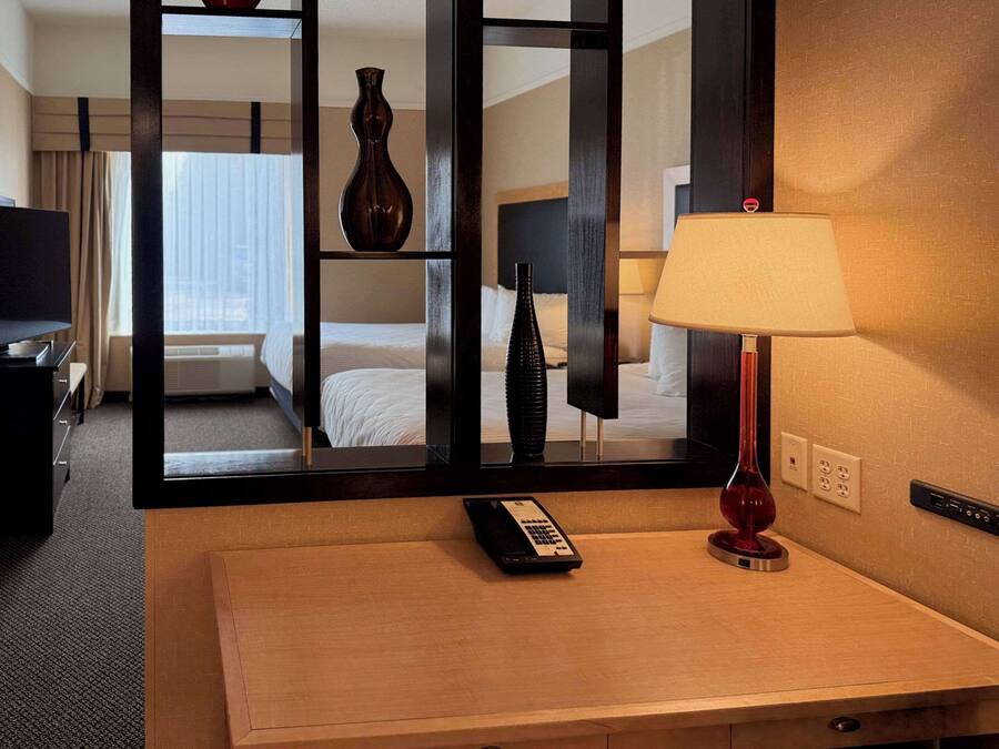A hotel room with a wooden desk, a remote control, a lamp, and a mirror reflecting the room.
