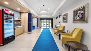 Un vestíbulo de hotel con refrigerador de Coca-Cola, alfombra azul, sillas amarillas y cuadros en la pared.