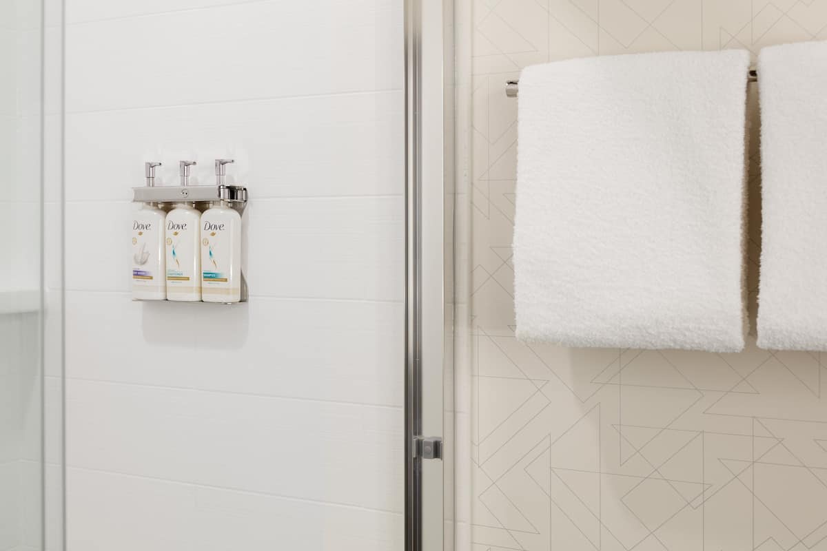 Un baño con pared de azulejos blancos, mampara de vidrio para la ducha y tres botellas de champú Dove montadas en la pared.