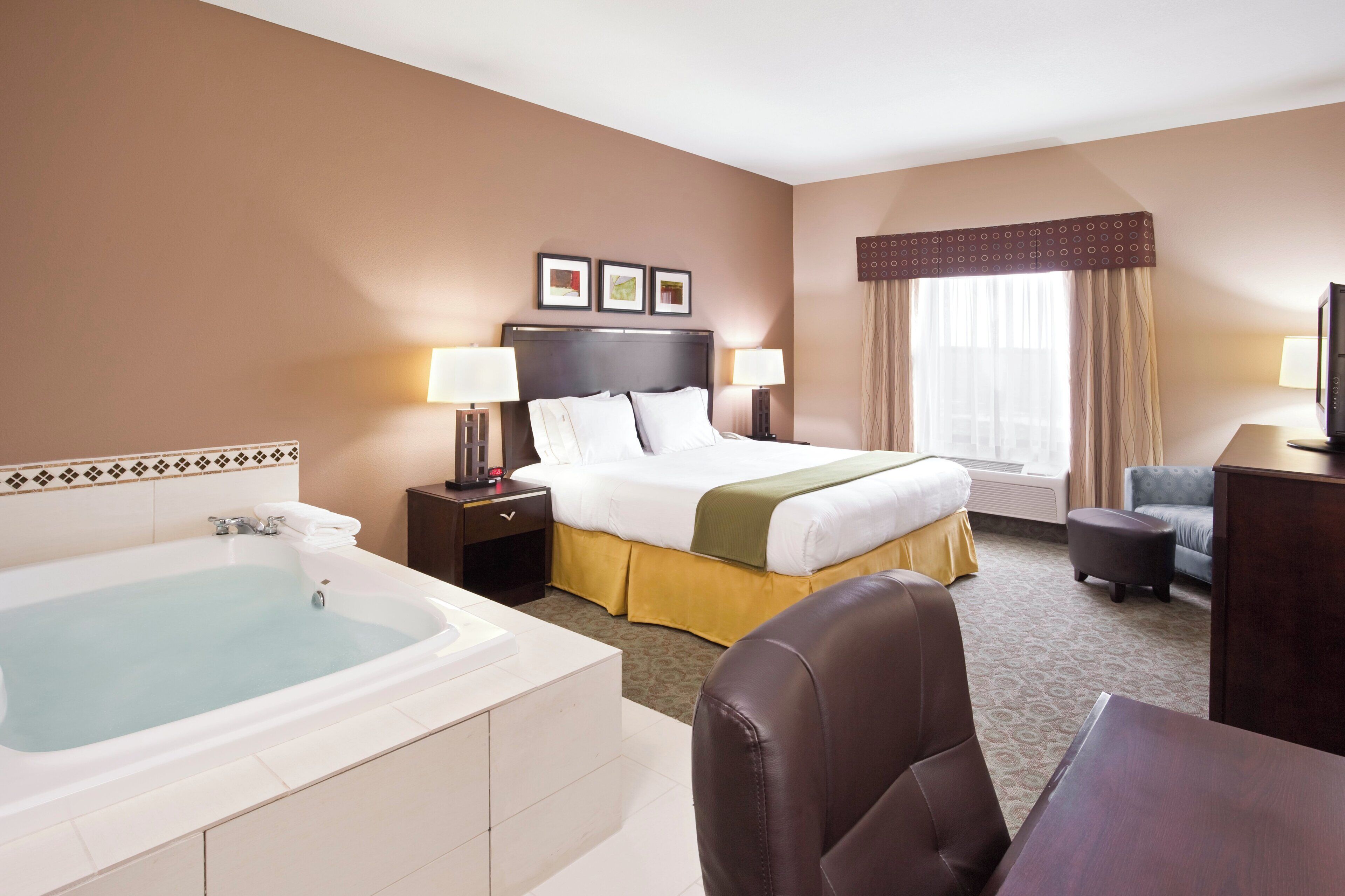 A hotel room with a large bed, a separate desk, a chair, a desk lamp, a bedside table with a lamp, a large bathroom with a bathtub, and a window with curtains.