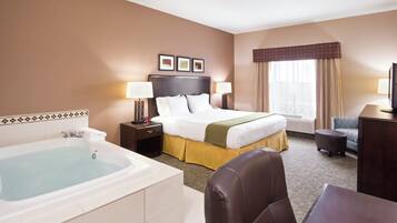 A hotel room with a large bed, a separate desk, a chair, a desk lamp, a bedside table with a lamp, a large bathroom with a bathtub, and a window with curtains.