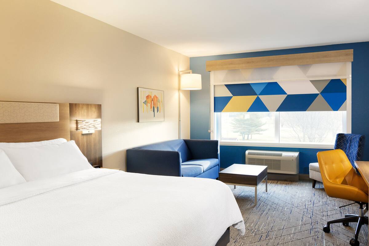 Habitación de hotel con cama, sofá, escritorio y silla. Se ve una ventana con vistas a árboles y una lámpara en la pared.