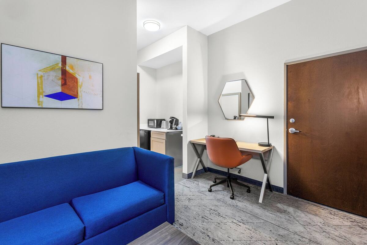Habitación de hotel moderna con un sofá azul, un escritorio con una silla naranja, una puerta de madera y un espejo grande en la pared.