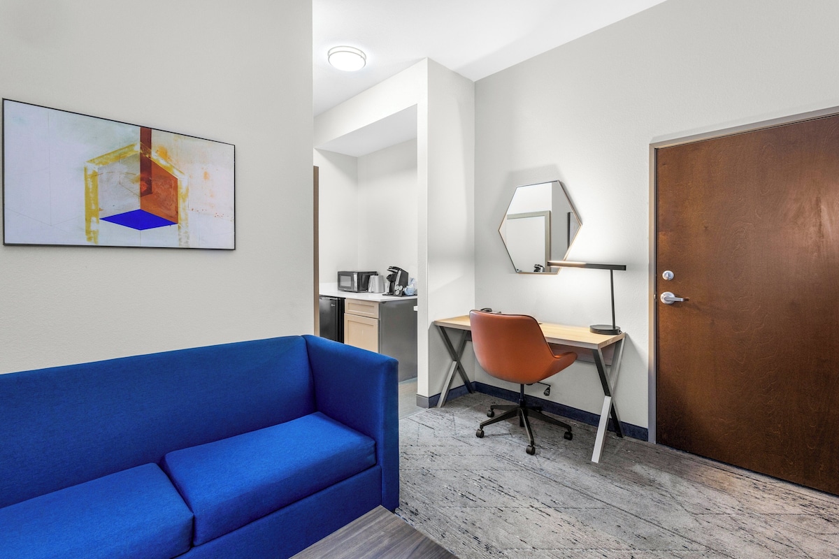 Una habitación de hotel con un sofá azul, un escritorio con una silla, un microondas, una cafetera y una puerta con espejo.