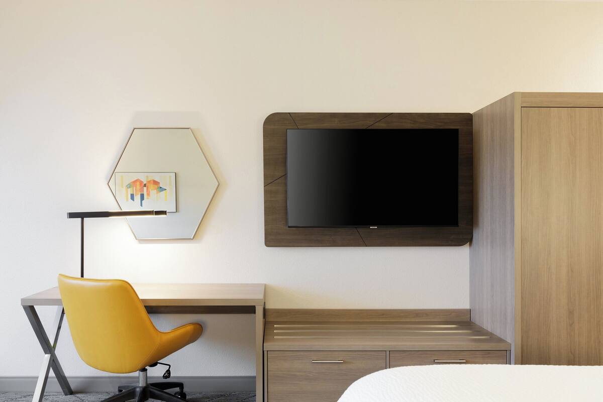 Una habitación de hotel con cama, escritorio con silla, televisor montado en la pared y armario de madera.
