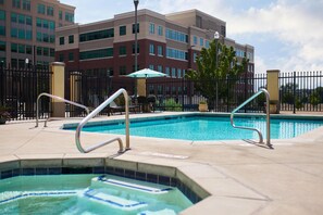 A swimming pool area with a hot tub, outdoor seating, and a building in the background.