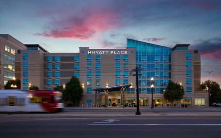 A modern Hyatt Place hotel with a prominent glass facade, a covered entrance, and a red bus in motion.